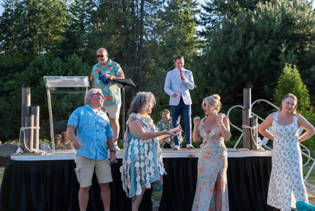 Outdoor stage event. Man speaks, woman wears oyster shell dress, others watch and react.