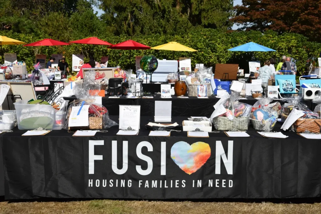 Outdoor fundraiser: FUSION banner on table packed with gift baskets under colorful umbrellas.