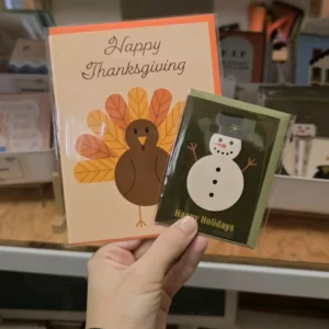 Hand holding a Happy Thanksgiving turkey card and a Happy Holidays snowman card.