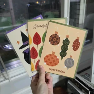 Hand holding three colorful seasonal cards: Halloween witch, autumn leaves, and holiday ornaments.