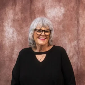 Joyful woman, grey hair, glasses, black top with heart necklace, against mottled background.