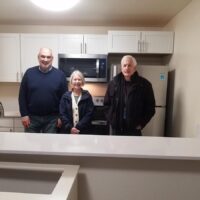 Three smiling older adults stand in a modern kitchen with white cabinets and new appliances.