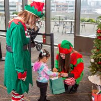 Two Christmas elves give a gift from a green bag to a small child.